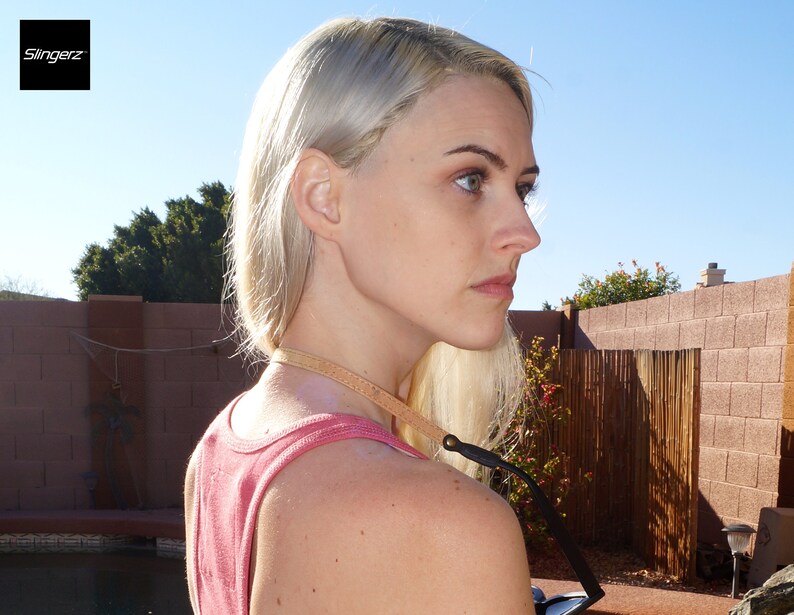 K&ouml;nnte beinhalten: Eine Frau mit blonden Haaren tr&auml;gt ein rosafarbenes Tanktop und eine braune Lederkameragurt. Der Gurt ist an einem schwarzen Kameraobjektiv befestigt.