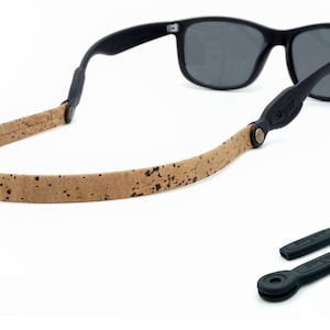 K&ouml;nnte beinhalten: Ein braunes Kork-Brillenband mit schwarzen Lederakzenten und Metallbeschl&auml;gen. Das Band ist an einer schwarzen Sonnenbrille mit dunklen Gl&auml;sern befestigt. Zwei schwarze Gummibrillenbandhalter sind ebenfalls enthalten.