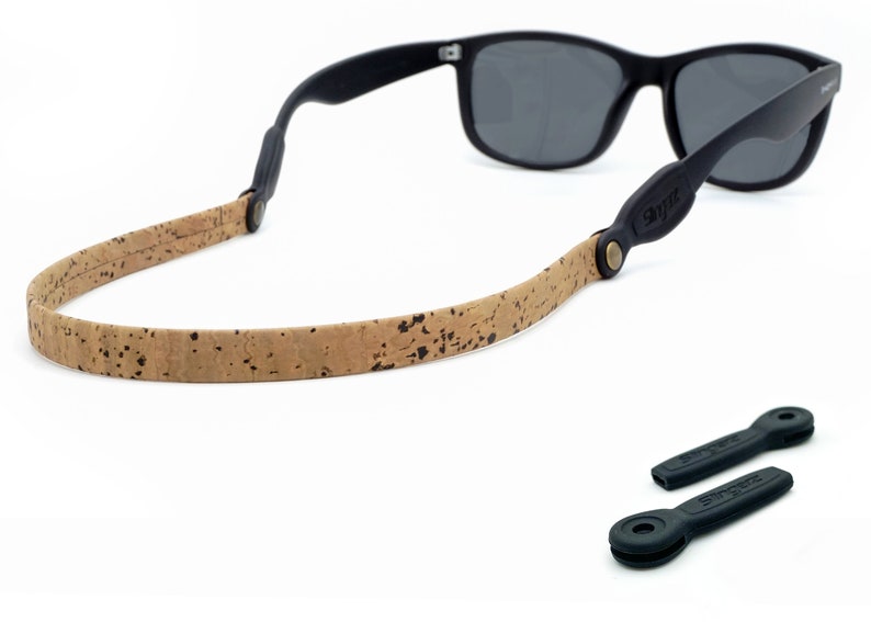 K&ouml;nnte beinhalten: Ein braunes Kork-Brillenband mit schwarzen Lederakzenten und Metallbeschl&auml;gen. Das Band ist an einer schwarzen Sonnenbrille mit dunklen Gl&auml;sern befestigt. Zwei schwarze Gummibrillenbandhalter sind ebenfalls enthalten.
