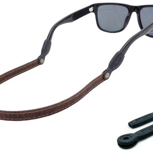 K&ouml;nnte beinhalten: Braunes Leder-Sonnenbrillenband mit N&auml;hten und schwarzen Gummieins&auml;tzen. Das Band ist an einer schwarzen Sonnenbrille mit dunklen Gl&auml;sern befestigt.