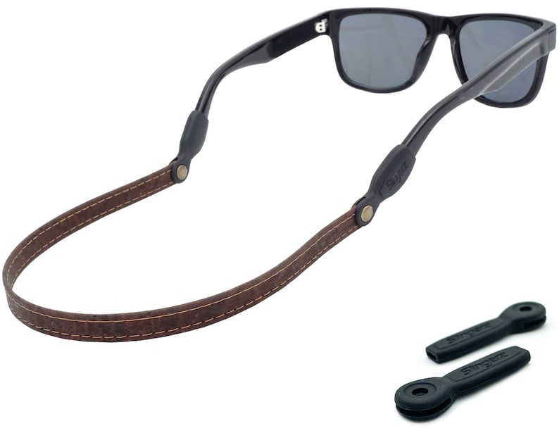 K&ouml;nnte beinhalten: Braunes Leder-Sonnenbrillenband mit N&auml;hten und schwarzen Gummieins&auml;tzen. Das Band ist an einer schwarzen Sonnenbrille mit dunklen Gl&auml;sern befestigt.