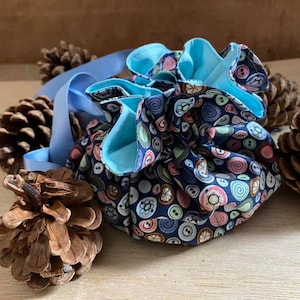 Puede incluir: Una bolsa redonda para joyas con cordón, con un exterior azul marino estampado con ilustraciones de botones coloridos. El interior es azul claro. Se adjunta una cinta azul claro. La bolsa está rodeada de piñas.