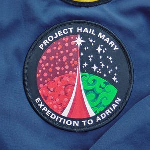 May include: A dark blue jacket featuring two circular embroidered patches. The top patch displays a spacecraft with a sunburst design and the text PROJECT HAIL MARY. The bottom patch shows a red and green planet with the text PROJECT HAIL MARY EXPEDITION TO ADRIAN.