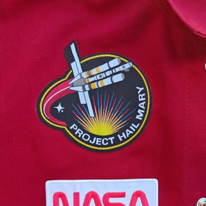 May include: Red jumpsuit featuring a NASA patch and a "Project Hail Mary" patch. The NASA patch has white lettering. The "Project Hail Mary" patch is circular with a rocket design and the text "PROJECT HAIL MARY".