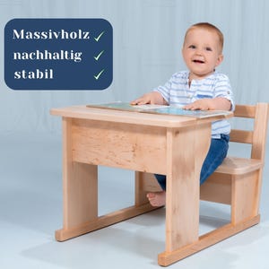 Può includere: Un set di scrivania e sedia per bambini in legno chiaro. La scrivania ha una superficie liscia e la sedia ha uno schienale e una seduta. Un bambino piccolo è seduto alla scrivania, guarda la telecamera e sorride. La scrivania è etichettata con le parole "Massivholz", "nachhaltig" e "stabil" con segni di spunta accanto a ciascuna parola.