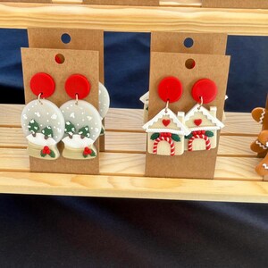 May include: A selection of festive Christmas earrings. The designs feature Santa Claus, snow globes, gingerbread houses, and gingerbread people. Each pair is attached to a brown card with a red circle. The earrings are displayed on a wooden shelf.