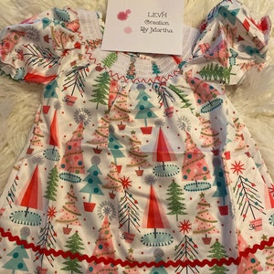Puede incluir: Un vestido blanco con un estampado de árboles de Navidad rojos y rosas. El vestido tiene mangas cortas y un dobladillo con volantes. El vestido está hecho por LEVH Creation by Martha.