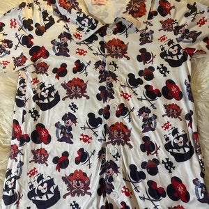 Pode incluir: Camisa branca de manga curta com botões e um padrão repetido de Mickey Mouse vestido de pirata, cabeças de Mickey Mouse e espadas e caveiras de piratas.