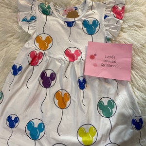 Puede incluir: Vestido de bebé blanco con un estampado de globos de Mickey Mouse de colores. El vestido tiene mangas con volantes y un cuello redondo. Una etiqueta rosa con texto es visible en el lado derecho del vestido.