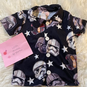 May include: Black short-sleeved button-up shirt with a white star and stormtrooper print. The shirt has a collar and buttons down the front.