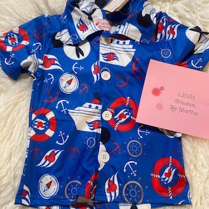 May include: A short-sleeved button-down shirt in blue featuring a nautical print with cruise ships, red and white life preservers, anchors, compasses, and ship wheels. A pink card with the text LEVH Creation By Martha is placed on the shirt.