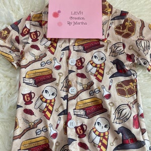 May include: A beige button-up shirt with a Harry Potter themed print. The print features owls, potions, glasses, keys, and other magical items. The shirt has a button closure and short sleeves.
