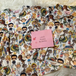 May include: Two white cotton dresses featuring a print of cartoon characters from the film Jurassic Park. The print includes dinosaurs, characters from the film, and a jeep. The dresses have short sleeves and a ruffled neckline. The dresses are on a white fluffy surface. The text on the pink card reads "LEVH Creation by Martha".
