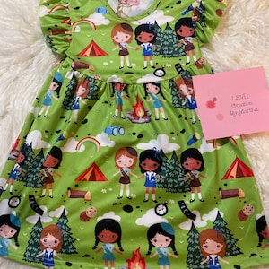 May include: A green sleeveless dress with a ruffled neckline and short sleeves. The dress features a pattern of girl scout figures, tents, trees, and other camping-related imagery. The text "LEVH Creation, by Martha" is printed on a pink card.