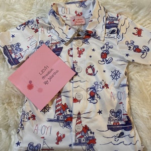 Op de afbeelding: Een wit, kort mouwig overhemd met knopen en een nautisch thema. Het overhemd heeft Mickey Mouse en Minnie Mouse figuren, vuurtorens, boten en de woorden "Ahoy!" in rood en blauw. Het overhemd heeft een label met de tekst "Levi Creations by Martha".