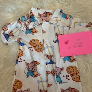 May include: White button-up shirt with a pattern of mice holding cookies and drinking milk. The shirt has a collar and short sleeves. The text "LEVH Creation By Martha" is on a pink card in the background.