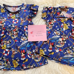 Pode incluir: Um vestido azul e branco e uma camisa azul e branca com um tema náutico com Mickey Mouse e seus amigos. O tecido tem um padrão repetido de Mickey Mouse, Minnie Mouse, Donald Duck, Pateta, Pluto e outros personagens da Disney. O vestido tem um decote com babados e mangas curtas. A camisa tem um colarinho e mangas curtas.