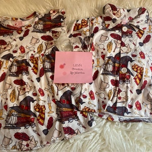 May include: A white dress and button-up shirt with a whimsical pattern featuring a wizard's hat, glasses, a cage, a feather, a key, a potion bottle, a candle, and a book. The fabric has a white background with red, yellow, and black accents. The text "LEVH Creation By Martha" is visible on a pink card.
