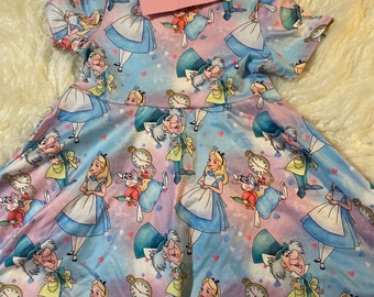 May include: A blue and pink dress with a whimsical Alice in Wonderland print featuring Alice, the Mad Hatter, and the White Rabbit. The dress has short sleeves and a full skirt.