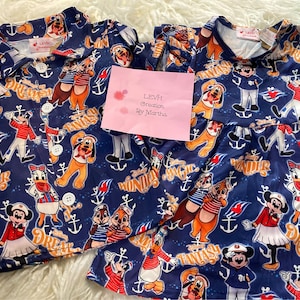 May include: A blue and white children's clothing set featuring a Disney Cruise Line theme. The set includes a short-sleeved button-down shirt and a dress with a ruffle trim. The fabric features a repeating pattern of Mickey Mouse, Donald Duck, Goofy, and other Disney characters. The text "Disney Wonder", "Disney Fantasy", and "Disney Dream" are also included in the pattern.