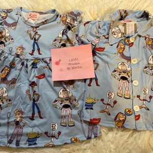 Pode incluir: Um conjunto de pijama azul e branco com tema Toy Story. O conjunto inclui um vestido de manga curta com acabamento de babados e uma camisa de botões. Os pijamas apresentam personagens como Woody, Buzz Lightyear e Jessie.