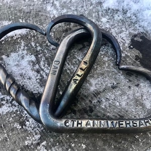 May include: Hand-forged metal heart-shaped sculpture. One heart is twisted, the other is smooth, both are interlocked. The sculpture is engraved with "DAN", "ALLI", and "6TH ANNIVERSARY". The metal has a dark, rustic finish.