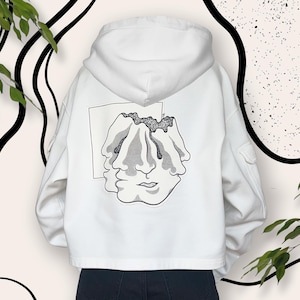 Op de afbeelding: Witte hoodie met een zwart-witte afbeelding van een gezicht op de rug. De afbeelding is van een gezicht met drie ogen en een mond.