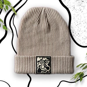 Op de afbeelding: Een beige gebreide beanie met een zwart-wit abstract gezichtontwerp aan de voorkant.