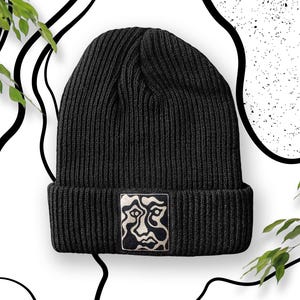 Op de afbeelding: Zwarte gebreide beanie met een zwarte en witte abstracte gezichtsontwerp patch.