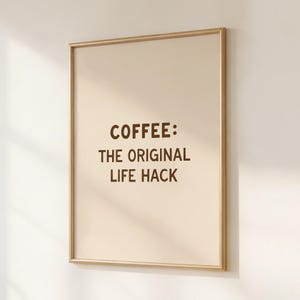 May include: A framed print with the text "COFFEE: THE ORIGINAL LIFE HACK" in brown lettering. The print has a beige background and a gold frame, displayed against a white wall. A simple, minimalist design.