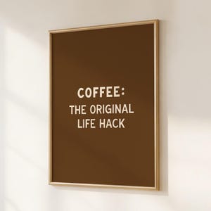 May include: A framed wall art piece with a brown background and gold frame. The white text reads "COFFEE: THE ORIGINAL LIFE HACK". The artwork is displayed on a white wall.