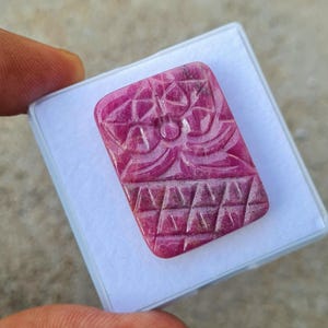 May include: A rectangular, deep pink carved gemstone featuring a stylized floral motif on the upper portion and a geometric grid pattern of triangles on the lower section. The stone has a smooth, polished surface and is set against a white background.