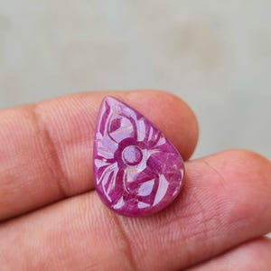 May include: A teardrop-shaped gemstone in a deep magenta-pink hue, featuring a carved floral pattern on its surface. The stone has a smooth, polished finish with visible natural inclusions and a distinct central circular detail within the petals.