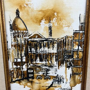 May include: A framed painting of a city scene in brown and white tones. The painting depicts a building with a dome and other buildings in the background. The artist's signature is visible in the bottom right corner.