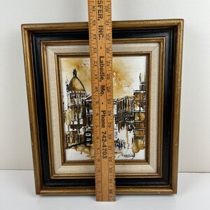 May include: A framed black and white painting of a city scene with a brown and white color scheme. The painting is in a gold and black frame.