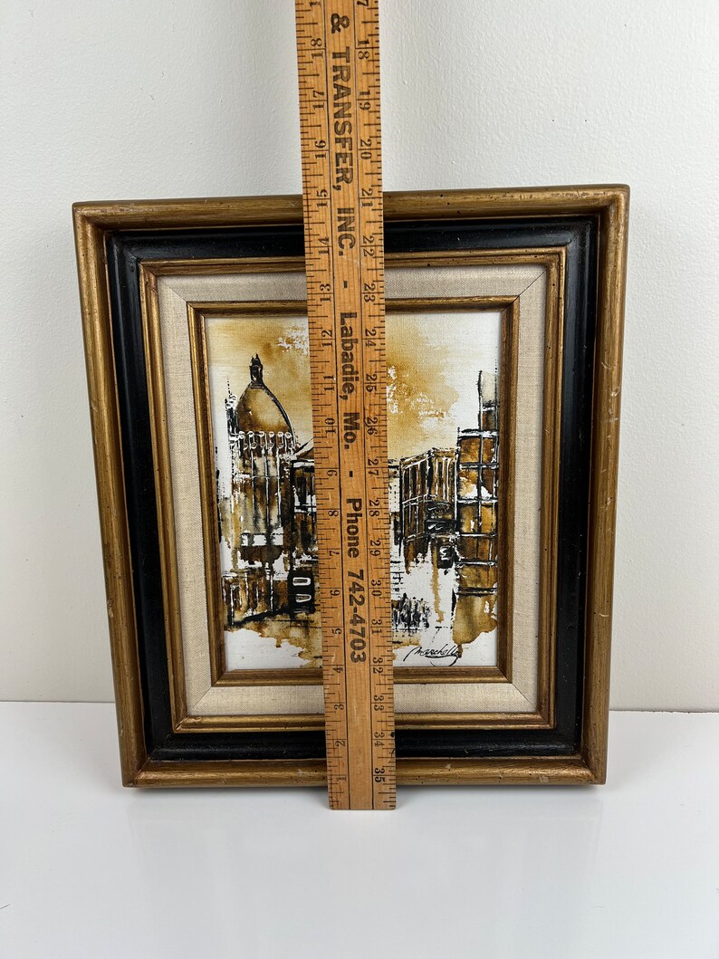 May include: A framed black and white painting of a city scene with a brown and white color scheme. The painting is in a gold and black frame.