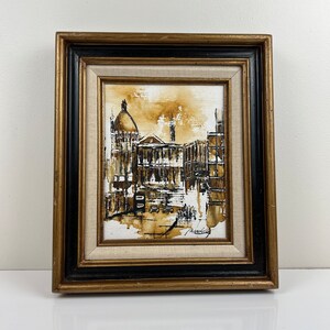 May include: A framed painting of a city scene in shades of brown and black. The painting depicts a building with a dome and other buildings in the background. The artist's signature is visible in the bottom right corner.