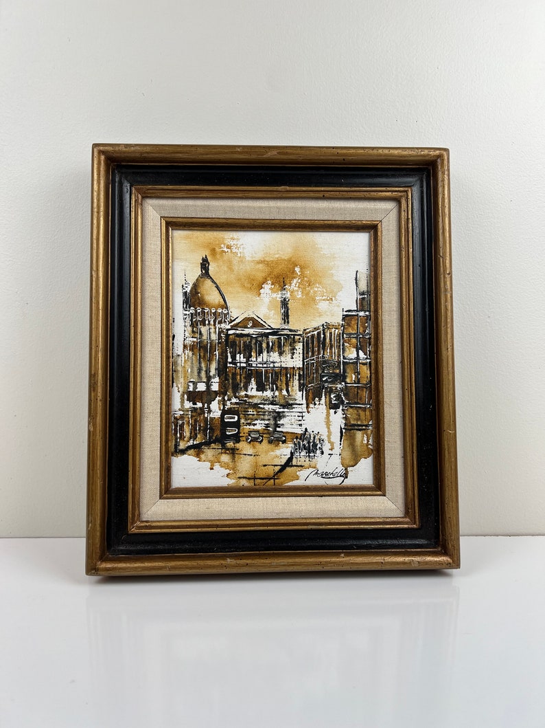 May include: A framed painting of a city scene in shades of brown and black. The painting depicts a building with a dome and other buildings in the background. The artist's signature is visible in the bottom right corner.