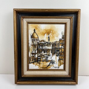 May include: A framed painting of a city scene in sepia tones. The painting depicts a building with a dome and other buildings in the background. The painting is signed in the bottom right corner.