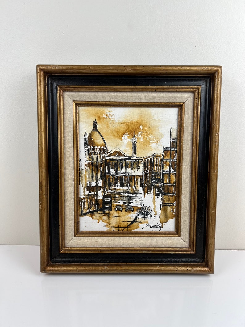 May include: A framed painting of a city scene in sepia tones. The painting depicts a building with a dome and other buildings in the background. The painting is signed in the bottom right corner.