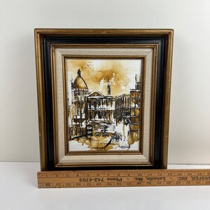 May include: A framed sepia-toned painting of a city street scene with buildings and a dome-shaped structure in the background. The painting is in a black and gold frame.