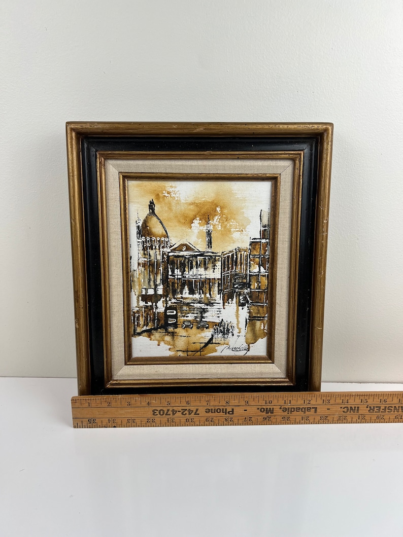 May include: A framed sepia-toned painting of a city street scene with buildings and a dome-shaped structure in the background. The painting is in a black and gold frame.