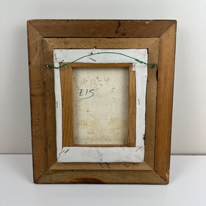 May include: The back of a framed painting with a wooden frame. The canvas is white and has the text "HA 12-44 E15" written on it.