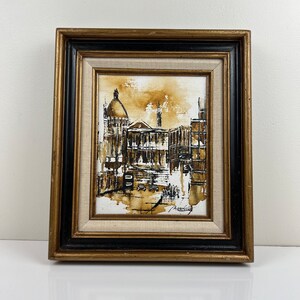 May include: A framed painting of a city street scene in shades of brown and black. The painting depicts a building with a dome and other buildings in the background. The artist's signature is visible in the bottom right corner.