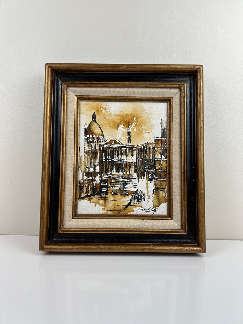 May include: A framed painting of a city street scene in shades of brown and black. The painting depicts a building with a dome and other buildings in the background. The artist's signature is visible in the bottom right corner.