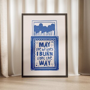 May include: A framed print with a blue illustration of a matchbox with the text "MAY the bridges I BURN light the WAY" inside. The background is a white wall with beige curtains.