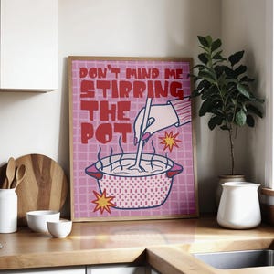 May include: A framed print with the text "DON'T MIND ME STIRRING THE POT" in red letters. The image features a pink pot with steam rising, a hand stirring, and starbursts. The print is displayed in a kitchen setting with white cabinets and a wooden countertop.