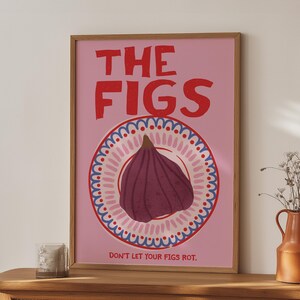 Könnte beinhalten: Ein gerahmtes rosa Poster mit den roten Worten "THE FIGS". Darunter eine Feigenillustration auf einem Teller mit blauem und weißem Rand. Der Text "DON'T LET YOUR FIGS ROT" steht unten. Das Poster steht auf einem Holztisch.