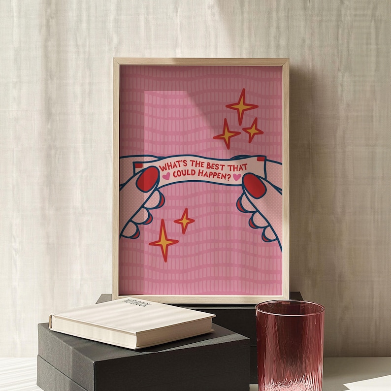 Peut inclure: Une affiche encadr&eacute;e avec un fond rose et le texte "WHAT'S THE BEST THAT COULD HAPPEN?" tenu par des mains illustr&eacute;es. L'affiche pr&eacute;sente des &eacute;toiles dor&eacute;es et un cadre en bois clair. Un carnet et un verre sont au premier plan.