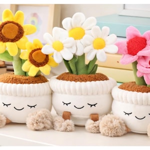 May include: Three plush flower pot toys with smiling faces. The flowers are yellow sunflowers, white daisies, and pink flowers. The pots are white with brown 'soil' and tan pom-pom feet.